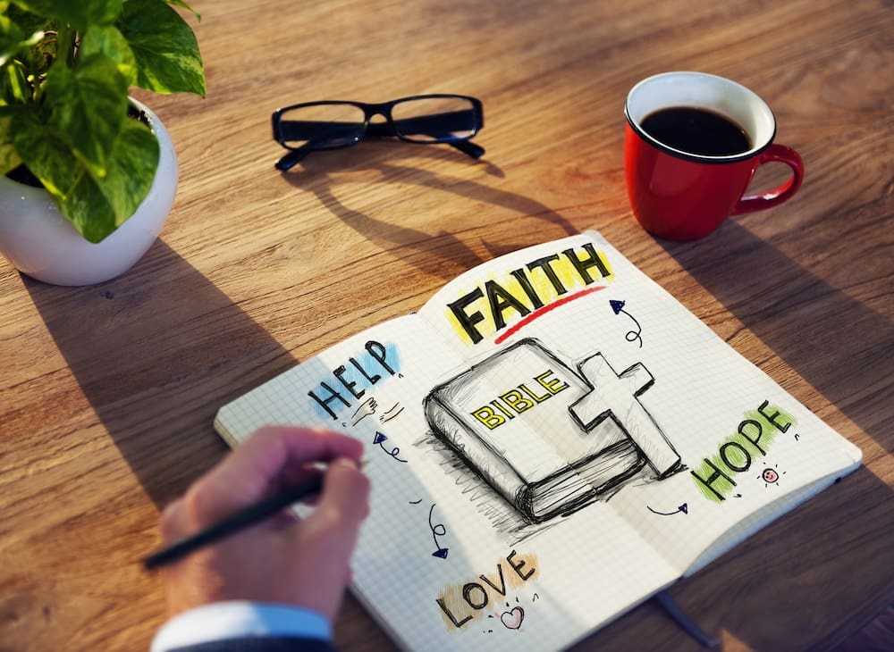 Image of a journal as seen from above, open to a page filled with illustrations of the Bible, cross, and the words 'faith', 'hope', and 'love'. The journal is surrounded by a full coffee cup and reading glasses.